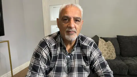 BBC Tariq Jahan at his home. He is wearing a checked shirt with glasses tucked in the front.