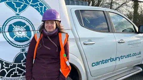 Canterbury Archaeological Trust Jess Twyman is standing next to a van which has the trust logo on it. She is wearing a burgundy jumper and hi-vis jacket and a purple hard hat. She has long hair and is smiling at the camera.