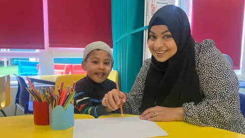 A boy in a grey hat and a stripy navy jumper sits beside a lady in a black and white dress and a black hijab. They are both holding an orange pencil and writing. They are in a classroom sitting at a yellow table with yellow seats and red blinds behind them