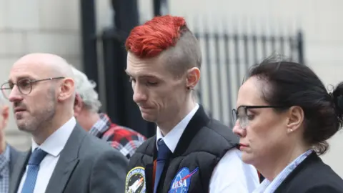 Pacemaker Mr Doyle is looking down to the ground with a sad expression. He has a his hair died red, is wearing a white shirt, black tie and black body warmer