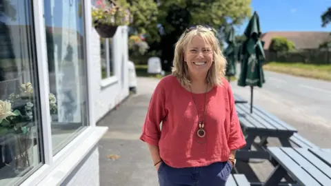 A woman with mid-length blonde hair with sunglasses on her head. She stands with her hands in her pockets and smiles at the camera. She is outside the pub and there are picnic tables and hanging baskets in the background.
