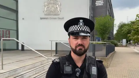 Ben Parker/BBC Sgt Chris Green wearing a black police uniform. He is standing in front of steps at Ipswich Crown Court with a large sign in the background, reading IPSWICH CROWN COURT.