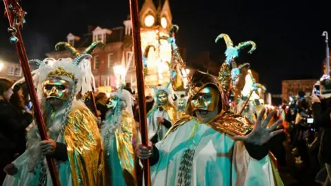 A group of people in white and gold with metallic mass covering most of their faces. The sky is dark and they are holding up long sticks with animals on them. There is a clock tower in the background which is lit up. The person at the forefront of the picture is looking into the camera and holding up their hand with their mouth open. There are crowds on either side of the people in costumes.