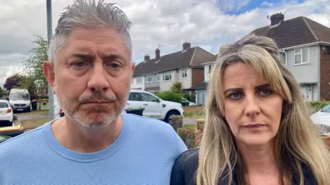 A sad looking couple, the man with grey hair and a blonde woman, are stood on a residential street.