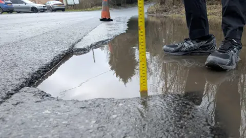 Someone wearing black and grey trainers sticks a yellow metal tape measure into a water-filled pothole. A single traffic cone stands nearby. Three cars are parked in front of a large metal gate on the opposite side of the road.