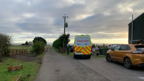 A police van stands at the end of a lane leading to the seaside