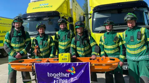 Six men are wearing green and yellow uniforms with helmets. They are standing in front of ambulances while holding an orange stretcher. 