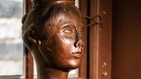 A close-up of a wooden carving of Queen Victoria, showing her as a young woman. The carving is detailed and reveals her hair pulled back in a bun. Its nose has been damaged. The figure is side on and has its back to a window