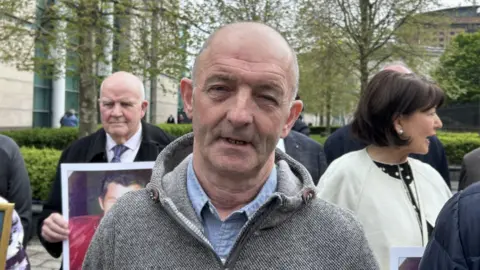 A bald man wearing a blue shirt and grey jumper. There is a group of people behind him. 