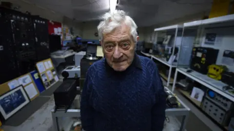 Pacemaker Fred Jennings, an elderly man with white hair wearing a knitted blue jumper, pictured in the Radio Room named after him at the Ulster Aviation Society's hangar at the former Maze prison site.