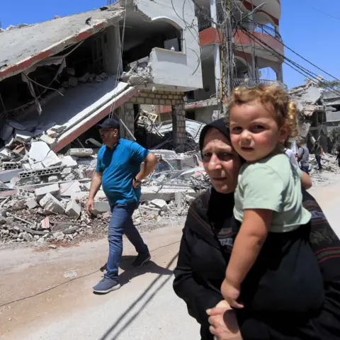 Getty Images A woman carries a small girl while walking down a street in front of a destroyed building 