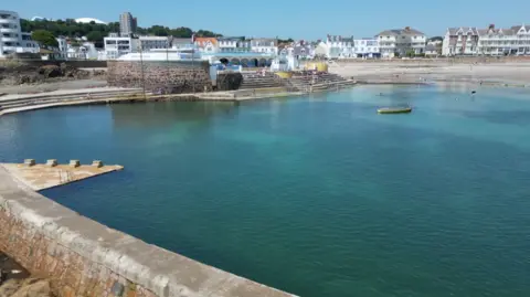 The lido at Havre des Pas. The water is clear and there are buildings with steps down to it. In the background are houses, two high rise buildings and a beach.