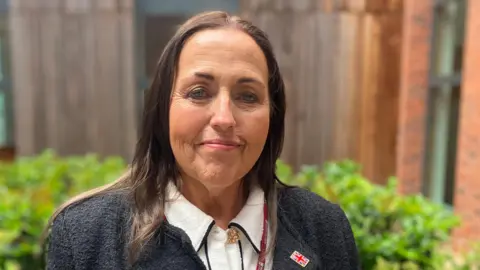 Councillor Joanne Monk photographed outside the council buildings. She has long brown hair and is wearing a light shirt, dark jacket and a lanyard. She has a Union Jack pin badge on her jacket lapel and is smiling at the camera. There is a low green bush behind her. 