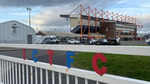A white gate with the letters I C T F C in red and blue writing in front of a brick football stand with orange pillars. Various cars are in the foreground of a car park, as well as a grassy area to the right of the picture. 