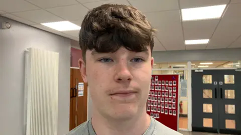 Owen is standing in a school hallway. He has brown hair which is quite long at the front and is wearing a grey top. Behind him a school corridor can be seen including a set of double doors and a tall radiator.