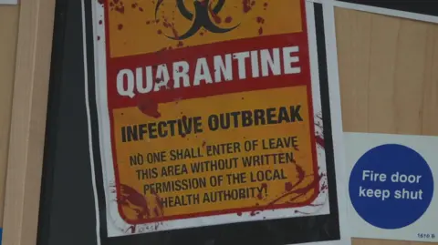 Big yellow quarantine sign on a classroom door. It reads: Infective Outbreak. No one shall enter or leave this area without the written permission of the local health authority! A sign behind it reads: Fire door. Keep shut.