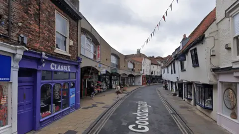 Google Goodramgate with shops on the left and right hand side, with a road going down the middle with double yellow lines on each side. 