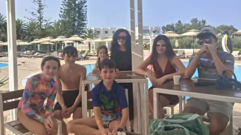 Georgina Doherty, una madre y seis niños de diferentes edades, están sentados en un chiringuito junto a la playa. Todos llevan traje de baño.