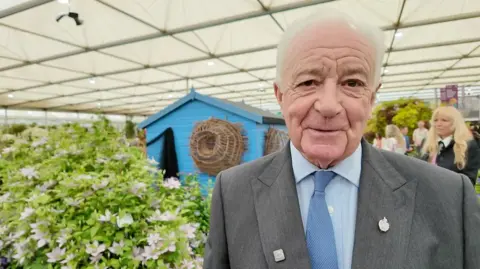 A man in a grey suit and blue shirt with a blue tie standing inside a tent at Chelsea Flower Show. There is a variety of plants and flowers, and a blue shed in the background.  
