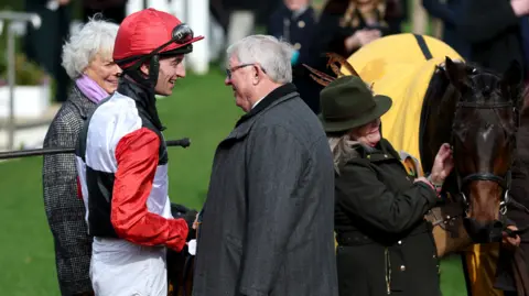 Action Images/Reuters Jockey Patrick Mullins shakes hands with former football manager Alex Ferguson. Mullins wears black and white silks with red sleeves and a red hat, while Ferguson wears a grey overcoat. Another horse and other punters are visible in the background.