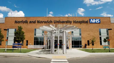 The refurbished outpatients' entrance at the Norfolk and Norwich University Hospital. It shows a white revolving door to the front, with signage to the left and some trees and bushes outside.