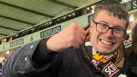 Daris Nesbitt Daris Nesbitt, who is wearing a navy jacket and a Scotland scarf, waves his right fist in the air as he celebrates the national team's 4-2 victory over Denmark at Hampden. He has short dark hair, dark-rimmed glasses and is grinning widely.
