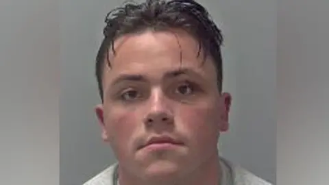 A custody picture of Simon Doherty, looking straight at the camera. He is wearing a grey top and has dark short hair, with some strands of hair hanging down his forehead