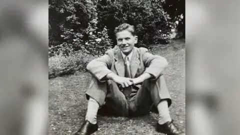 A black and white picture of Gordon wearing a light shirt, tie and smart trousers with a light coloured over blazer.  He is wearing black shoes and light socks and is smiling at the camera, with trees behind him. It was taken in his garden in 1951. 