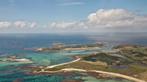 Isaac Ogden A drone photo across the smaller islands in the Isles of Scilly. The water is clear and shades of blue, green and turquoise and the beaches lining the islands are white. There is a blue sky and some white clouds.