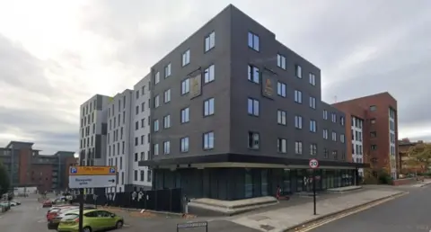 Google Maps A large grey building with numerous windows and a glass-fronted entry way.