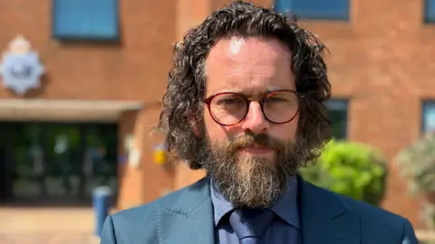 Jo Black/BBC A detective inspector stands outside police HQ, he has long dark hair and a beard. He has red-rimmed glasses.