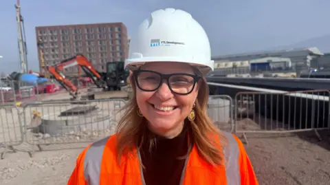 Helen Godwin is wearing a white hard hat and high vis jacket. She is pictured on a building site, smiling at the camera.
