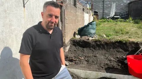 BBC Tom Clarke, who has dark hair with silver speckles and a dark beard, smiles at the camera. He wears a black polo shirt and white shorts and his dug up garden can be seen in the background. 