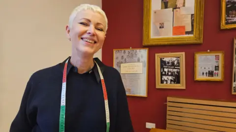 Aisha Iqbal/BBC A woman with cropped blond hair stands smiling, with a tailor's tape measure hung around her neck.