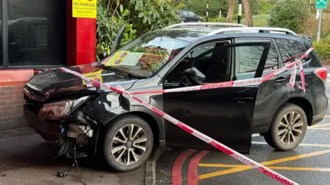 Eddie Mitchell A black MPV with a damaged front sits hall on a curb in front of a building, A yellow sign reads "no parking" and the back half of the car is on a box junction. It is cordoned off with tape 