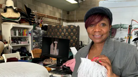 Helen Miller A woman wearing a grey jacket holding a piece of white fabric in her left hand. She is smiling as she looks into the camera. She is standing in a studio with hats in the background.