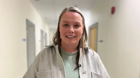Vickie Underhill stands in a hospital corridor and smiles at the camera. She is wearing a grey jumper with a lighter grey jacket over the top of it. She has shoulder-length fair hair