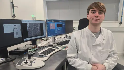 A bank of computer screens showing microscopic images. To the right is a younger man wearing a white lab coat