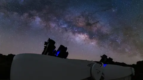 A night sky over several telescopes, black cylinders pointing towards the sky. The sky is brightly lit with stars and ranges in colours from deep blue to white.