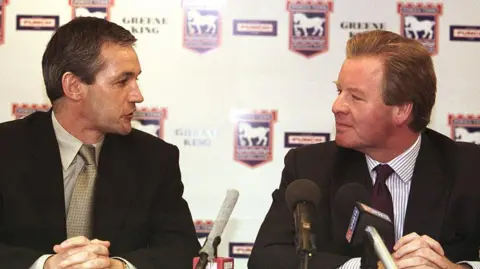 Getty Images Two men sit next to each other behind a desk at a football press conference. The man on the left has dark hair and looks over toward the other man as he talks. He has his hands resting in one another on the desk in front of him. He wears a black suit, grey shirt and tie. There is a microphone between the men. The man on the right also has dark hair and is looking toward the other man as he listens to him talking. He also wears a black suit with a white shirt with blue stripes on it and a dark purple tie. He also has his hands resting together on the desk in front of him.