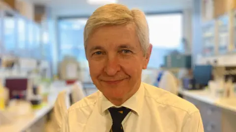 Sir John Burn, Professor of Clinical Genetics is in a laboratory in Newcastle. Looking straight at the camera and smiling, he is wearing a while shirt and dark tie.
