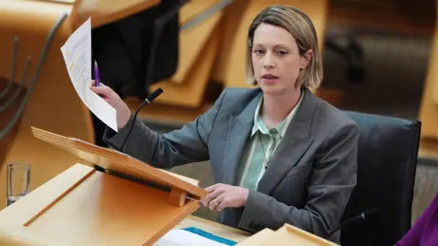 PA Media Jenny Gilruth, who has shoulder-length blonde hair and is wearing a grey jacket and a green shirt sits in the Scottish Parliament, holding a piece of paper