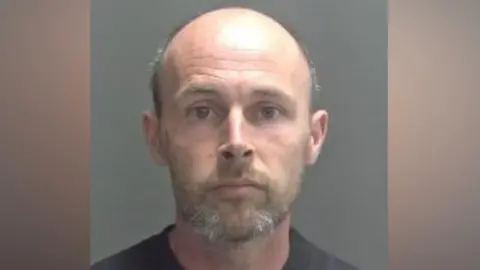 A police custody photo of Hudson. He has a short brown-and-grey beard and moustache and is bald. He is wearing a black T-shirt and looking into the camera.