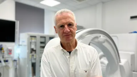 Tom Binet, a man with short white hair, is wearing a white shirt and no tie. He is standing in front of medical equipment, in a room which appears to be in a hospital. He is staring directly at the camera.