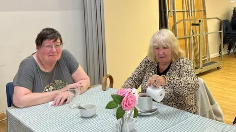 Two women are sitting on a green gingham table clothed table and are drinking coffee. They are sitting in a community hall with stacked tables behind them. 