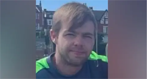 Man with brown hair and long fringe - he is wearing a grey t-shirt with neon lapels  - sitting in front of houses 