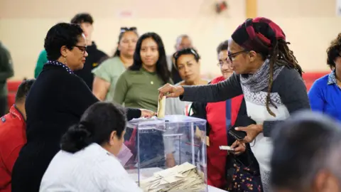 Getty Images A woman with a red hair covering places her vote in a see-through ballot box - a line of people can be seen queuing up to vote behind her in the French Pacific territory of New Caledonia