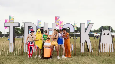 Josh Collins/Truck Festival A group of seven young men stand in front of a large letter display in a field. The letters read 'Truck' and there is the Truck Festival logo and multicoloured festival flags in the backgrounds. From left to right the men are wearing various costumes. The first man is topless with a Union Jack top hat and matching trousers, with a stachel bag strap across his torso, next to him a man stands wearing a banana costume, the next man wears a green tank top with yellow writing which says 'Jamaica', another man is crouched on the ground which his arms crossed dressed like an orange and white traffic cone. Then standing is a man who is topless wearing golden shorts and a gold bucket hat, a man wearing a blue and white cheerleader's costume and a man in a 70s style outfit with a black afro wig.