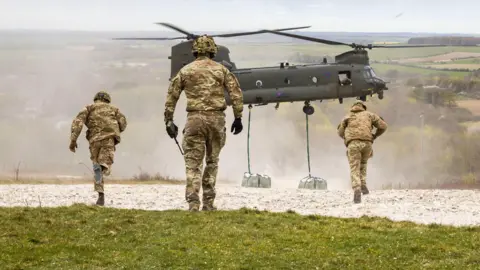 Soldiers walk towards a helicopter which is dropping white bags onto a chalk surface on a hillside