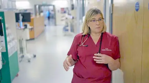 Consultant hepatologist Debbie Shawcross wearing red scrubs and a stethoscope around her neck, standing in a hospital corridor.